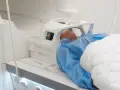 Pacjent w niebieskim fartuchu leży w aparacie do rezonansu magnetycznego. Czas badania, czyli ile trwa rezonans głowy, zależy od jego celu.