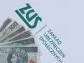 Pieniądze, w tym banknoty 100 i 200 zł, leżą obok zielonego logo ZUS. To dodatek dopełniający do świadczeń.
