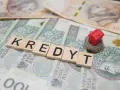 Litery układające słowo "KREDYT" na banknotach. Mały czerwony domek na stosie monet symbolizuje pomoc w spłacie kredytu, np. z funduszu wsparcia kredytobiorców.