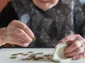 Dłoń starszej kobiety z pierścionkiem wkłada monetę do portmonetki. Rozrzucone monety na stole sugerują trudności finansowe, być może związane z emeryturą po mężu.