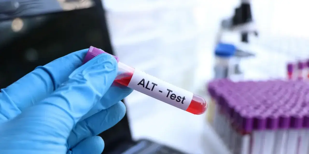 Ręka w niebieskiej rękawiczce trzyma probówkę z krwią oznaczoną "ALT - Test". W tle mikroskop i stojak z próbkami. To badanie ALT.