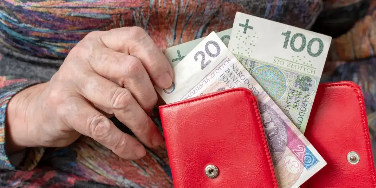 Dłoń starszej osoby wkłada banknoty 20 i 100 zł do czerwonego portfela. Czerwcowe emerytury wreszcie dotarły.