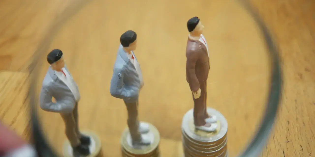Figurki mężczyzn w garniturach stoją na stosach monet. Jedna z nich jest wyższa, symbolizując wyższe zarobki, podczas gdy pozostałe są niższe, odzwierciedlając średnią krajową.