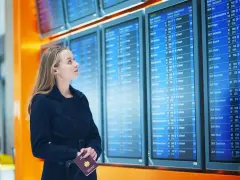 Kobieta z paszportem w dłoni sprawdza tablicę odlotów, przygotowując się do kontroli granicznej.