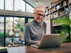 Uśmiechnięta kobieta z krótkimi blond włosami i okularami pracuje przy laptopie. Może sprawdza swój dodatek do wynagrodzenia.