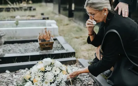 Kobieta w żałobie klęczy przy grobie, ocierając łzy, obok wiązanki kwiatów.