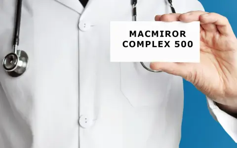 Lekarz w białym kitlu trzyma kartkę z napisem MACMIROR COMPLEX 500. Stetoskop wisi na jego szyi.