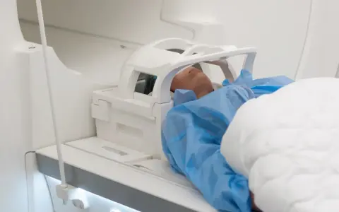 Pacjent w niebieskim fartuchu leży w aparacie do rezonansu magnetycznego. Czas badania, czyli ile trwa rezonans głowy, zależy od jego celu.