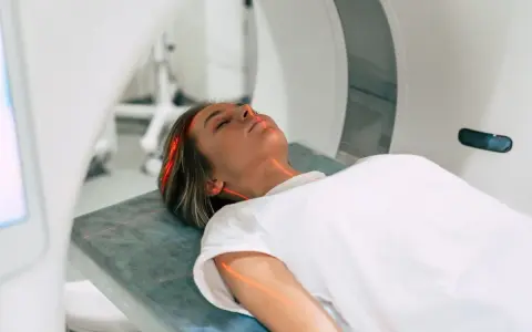 Kobieta leży na łóżku w aparacie do rezonansu magnetycznego. Czerwone linie lasera wskazują obszar badania. To właśnie mri co to za badanie?