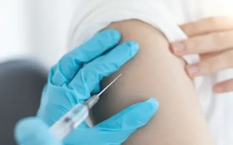 Ręka w niebieskiej rękawiczce podaje szczepionkę na kleszcze. Zabezpiecz się przed boreliozą i kleszczowym zapaleniem mózgu.