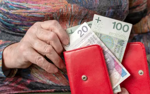 Dłoń starszej osoby wkłada banknoty 20 i 100 zł do czerwonego portfela. Czerwcowe emerytury wreszcie dotarły.