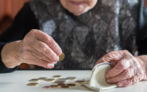 Dłoń starszej kobiety z pierścionkiem wkłada monetę do portmonetki. Rozrzucone monety na stole sugerują trudności finansowe, być może związane z emeryturą po mężu.
