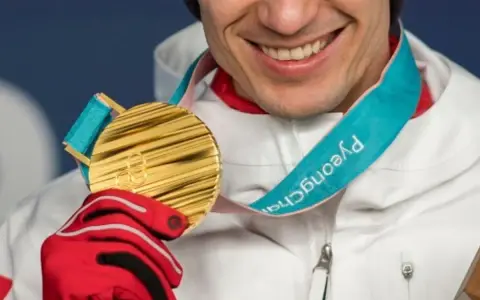Uśmiechnięty sportowiec w czerwonej rękawicy trzyma złoty medal olimpijski z PyeongChang. Taki medal to marzenie o emeryturze.