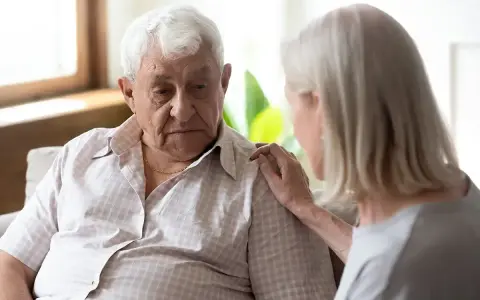 Starszy mężczyzna z zamyśloną miną, kobieta kładzie mu dłoń na ramieniu. Może to być wsparcie w trudnych chwilach, gdy pojawiają się psychoza objawy.