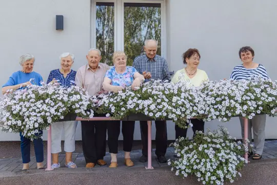 Siedmioro seniorów, otoczonych kwitnącymi petuniami, cieszy się słońcem. Wśród nich jest pani, która wygląda jakby właśnie sięgnęła po clemastinum.