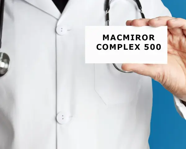 Lekarz w białym kitlu trzyma kartkę z napisem MACMIROR COMPLEX 500. Stetoskop wisi na jego szyi.