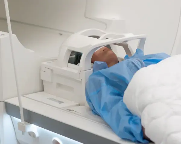 Pacjent w niebieskim fartuchu leży w aparacie do rezonansu magnetycznego. Czas badania, czyli ile trwa rezonans głowy, zależy od jego celu.