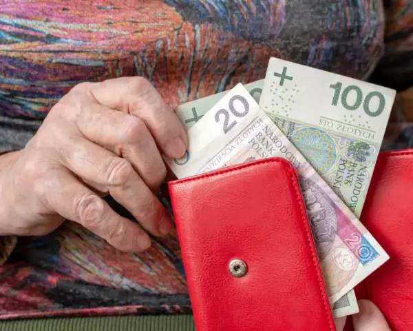 Dłoń starszej osoby wkłada banknoty 20 i 100 zł do czerwonego portfela. Czerwcowe emerytury wreszcie dotarły.