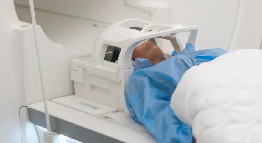 Pacjent w niebieskim fartuchu leży w aparacie do rezonansu magnetycznego. Czas badania, czyli ile trwa rezonans głowy, zależy od jego celu.