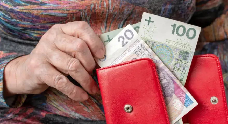 Dłoń starszej osoby wkłada banknoty 20 i 100 zł do czerwonego portfela. Czerwcowe emerytury wreszcie dotarły.