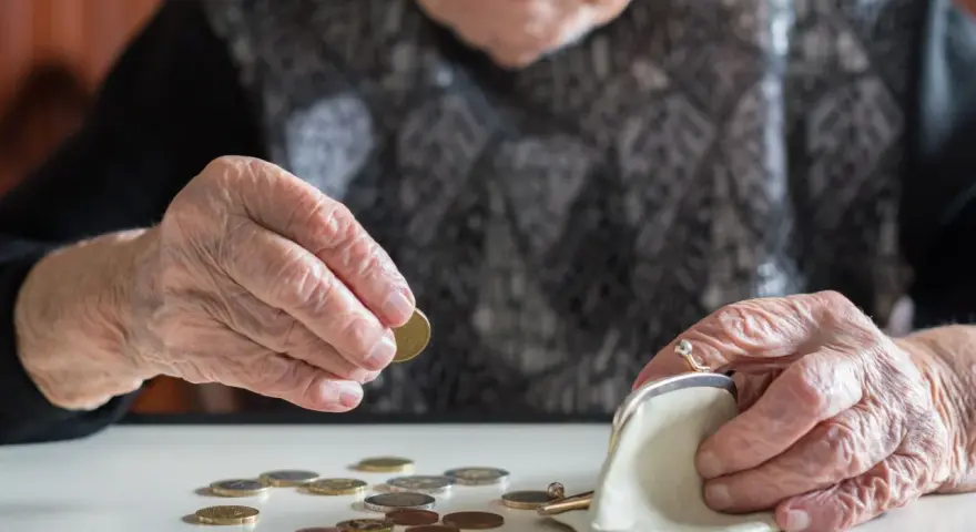 Dłoń starszej kobiety z pierścionkiem wkłada monetę do portmonetki. Rozrzucone monety na stole sugerują trudności finansowe, być może związane z emeryturą po mężu.