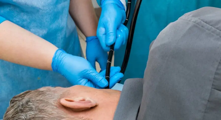 Lekarz w niebieskich rękawiczkach wprowadza endoskop. Pacjent leży na boku, przygotowany do gastroskopii.