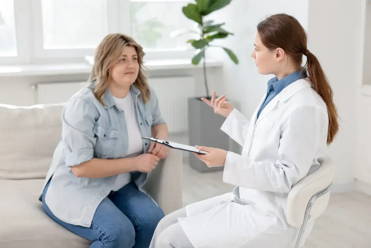 Pacjentka rozmawia z obesitologiem, który notuje informacje na kartce. Lekarz w białym kitlu wyjaśnia coś pacjentce siedzącej na kanapie.