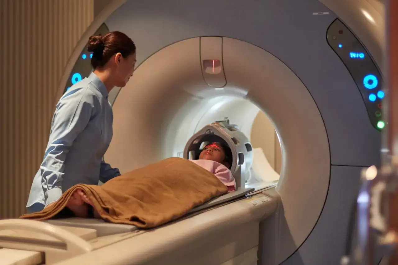 Dziecko leży w aparacie MRI, a pielęgniarka czuwa obok. To badanie, mri co to, pozwala zajrzeć do wnętrza ciała.