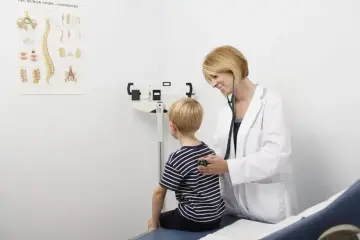 Lekarka osłuchuje chłopca podczas bilansu dziecka. W tle waga lekarska i plakat z anatomią kręgosłupa.